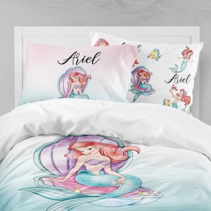Op de afbeelding: Een wit en roze kussensloop met een aquarel-illustratie van Ariel de zeemeermin die op een schelp zit. Het kussensloop heeft de naam "Ariel" in cursief erop.