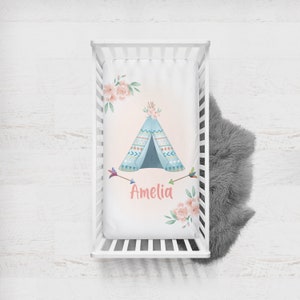 May include: A white crib sheet with a pink and blue teepee design, flowers, and arrows. The name Amelia is written in pink below the teepee.