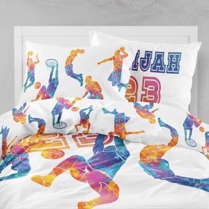 Puede incluir: Una funda nórdica blanca con un diseño de acuarela colorido de jugadores de baloncesto. El diseño presenta siluetas de jugadores en varias poses, incluyendo driblar, tirar y saltar. La funda nórdica también incluye el nombre "MALIA" y el número "23" en una fuente colorida y estilizada.