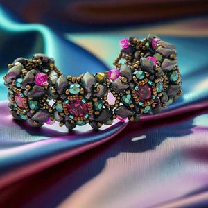Puede incluir: Una pulsera de cuentas con una base gris oscuro, acentuada con cuentas turquesas, rosas y doradas. La pulsera presenta grandes piedras preciosas rosas redondas y está sobre un fondo de tela brillante azul y morada.