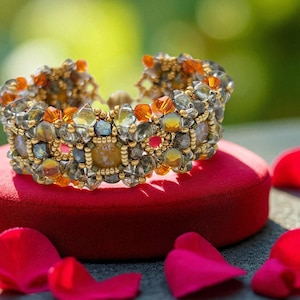 Puede incluir: Un brazalete de cuentas con una mezcla de cuentas transparentes, naranjas y doradas. El brazalete descansa sobre un cojín de terciopelo rojo, con pétalos de rosa rojos esparcidos alrededor. Es una joya.