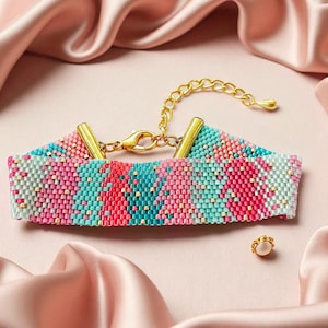 Puede incluir: Una pulsera de cuentas con un cierre y cadena dorados, con un patrón de cuentas rosas, turquesas y blancas. Un pequeño pendiente de botón dorado con una perla está cerca. El fondo es de satén rosa.