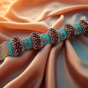 Puede incluir: Una pulsera de cuentas con un cierre dorado y un diseño central de cuentas turquesas y doradas alternas, con racimos de cuentas rojas. La pulsera se exhibe sobre una tela de satén color melocotón.