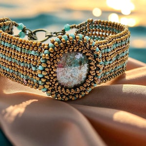 Puede incluir: Una pulsera de cuentas con cuentas doradas y turquesas. La pulsera presenta una piedra central grande y un cierre de color bronce. El intrincado trabajo de cuentas crea un diseño texturizado y llamativo. La pulsera se exhibe sobre una superficie satinada.