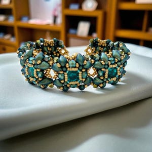 Puede incluir: Un brazalete de cuentas de color verde azulado y dorado. El brazalete presenta un patrón repetitivo de cuentas de color verde azulado y detalles dorados. Se muestra sobre una superficie de satén gris claro. El brazalete es una joya. Circunferencia de unos 17 cm.