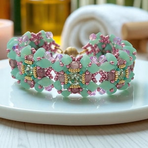 Puede incluir: Un brazalete de cuentas con una mezcla de cuentas verde menta, rosa y doradas. El brazalete está sobre un plato blanco. Las cuentas están dispuestas en un patrón geométrico.