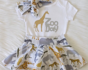 May include: A white baby onesie with a gold giraffe and the text "Zoo Day" in grey. The onesie is paired with a grey and white headband with a giraffe print and a white skirt with a multi-coloured animal print.