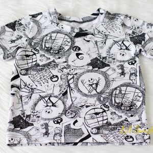 May include: White short-sleeved t-shirt featuring a black and white print of characters from the film The Nightmare Before Christmas. The print includes Jack Skellington, Sally, and other characters from the film.