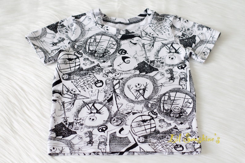 May include: White short-sleeved t-shirt featuring a black and white print of characters from the film The Nightmare Before Christmas. The print includes Jack Skellington, Sally, and other characters from the film.