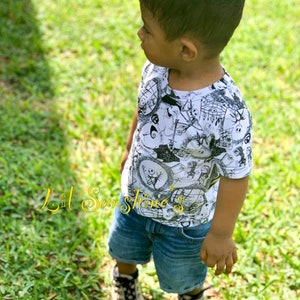 May include: A young child wearing a white t-shirt with a black and white print of characters from the film "The Nightmare Before Christmas". The child is wearing blue denim shorts and black and white trainers. The child is standing in a grassy area.