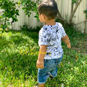 May include: A young child wearing a white t-shirt with a black and white graphic print and blue denim shorts. The child is standing in a grassy area. The text "Lil Sunshine's" is visible in the lower left corner of the image.