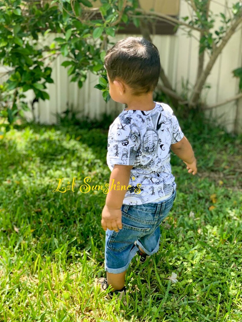 May include: A young child wearing a white t-shirt with a black and white graphic print and blue denim shorts. The child is standing in a grassy area. The text "Lil Sunshine's" is visible in the lower left corner of the image.
