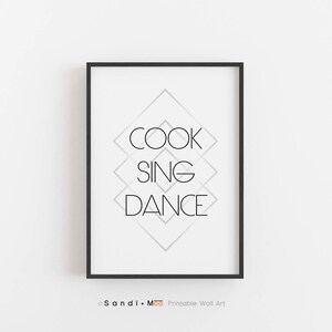May include: A framed print featuring the words "COOK SING DANCE" in a minimalist black font. The words are stacked vertically, set against a geometric diamond pattern. The frame is black, and the background is white.