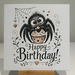 May include: A square greeting card featuring a black, fuzzy spider with large eyes holding a cupcake. The text reads Happy Birthday! in black script. The design includes a spiderweb, small hearts, and stars in black and orange ink on white.