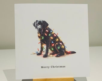 Puede incluir: Tarjeta de Navidad con un Labrador negro adornado con luces de colores. El perro está sentado, y las palabras "Merry Christmas" están impresas debajo. La tarjeta se muestra en un pequeño caballete de madera.