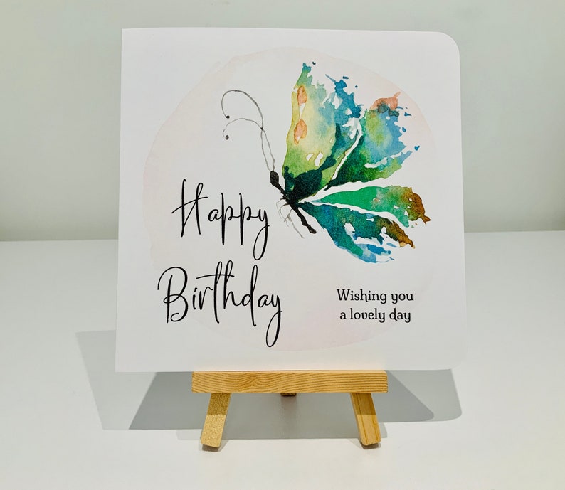Puede incluir: Una tarjeta blanca con las palabras "Happy Birthday" escritas en cursiva negra. Una pintura de acuarela de una mariposa verde y azul est&aacute; en la esquina superior derecha de la tarjeta. El texto "Wishing you a lovely day" est&aacute; escrito en negro debajo de la mariposa.