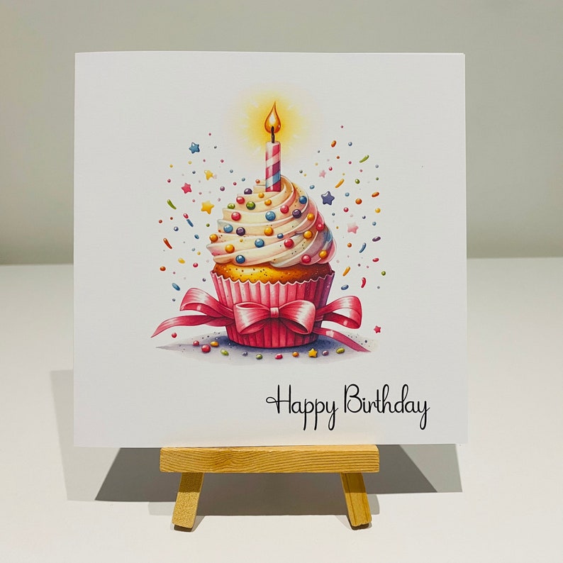 May include: A birthday greeting card featuring a watercolor illustration of a cupcake with a lit candle. The cupcake has white frosting, colorful sprinkles, and a pink bow. The words "Happy Birthday" are written in script.