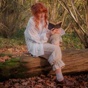 Puede incluir: Una mujer con el pelo largo y rojo se sienta en un tronco caído en un bosque, leyendo un libro. Lleva una camisa blanca y pantalones beige. El suelo del bosque está cubierto de hojas caídas.