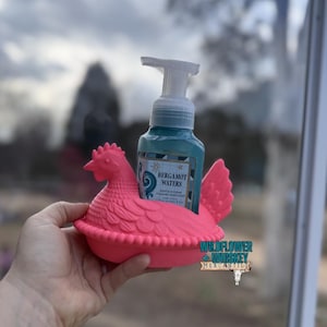 Puede incluir: Un jabonero con forma de gallina rosa brillante sostiene un dispensador de jab&oacute;n turquesa. El dispensador tiene una bomba blanca y una etiqueta que dice "Bergamot Waters". El soporte es de pl&aacute;stico y tiene un dise&ntilde;o detallado.