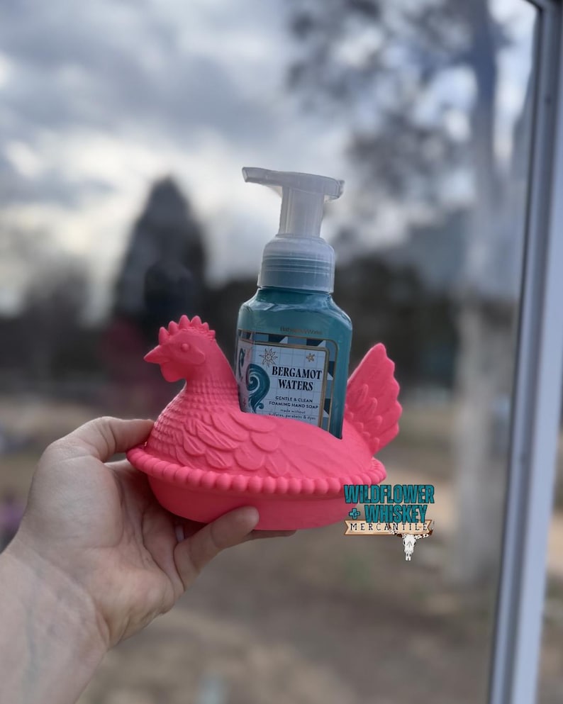 Puede incluir: Un jabonero con forma de gallina rosa brillante sostiene un dispensador de jab&oacute;n turquesa. El dispensador tiene una bomba blanca y una etiqueta que dice "Bergamot Waters". El soporte es de pl&aacute;stico y tiene un dise&ntilde;o detallado.