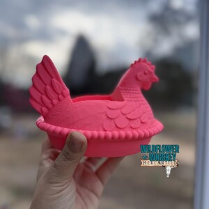 Puede incluir: Un recipiente decorativo rosa brillante con forma de gallina. El recipiente tiene un dise&ntilde;o detallado, que incluye una cresta, alas y una base con volantes. El texto "WILDFLOWER + WHISKEY MERCANTILE" es visible en el lateral.