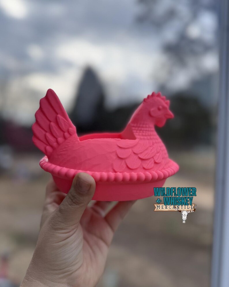 Puede incluir: Un recipiente decorativo rosa brillante con forma de gallina. El recipiente tiene un dise&ntilde;o detallado, que incluye una cresta, alas y una base con volantes. El texto "WILDFLOWER + WHISKEY MERCANTILE" es visible en el lateral.