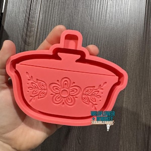 May include: A coral-colored silicone mold in the shape of a casserole dish. The mold features a floral design with a central flower and leaf accents. The mold has a handle on top and is held in a hand.