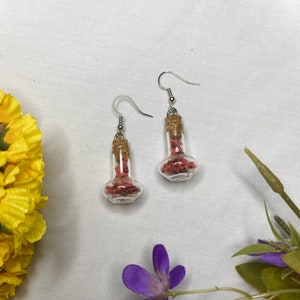 May include: A pair of silver earrings with small glass bottles filled with pink confetti. The bottles have cork stoppers.