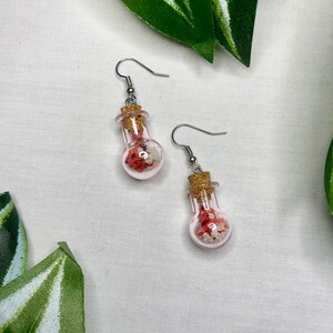 May include: A pair of earrings featuring small glass bottles filled with pink and white sand, each bottle has a cork stopper and hangs from a silver hook.