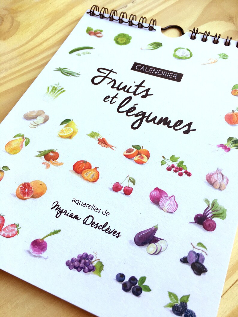 May include: A white calendar with a spiral binding. The calendar features a watercolor illustration of various fruits and vegetables, including strawberries, grapes, cherries, onions, and pumpkins. The text "Fruits et l&eacute;gumes" is written in black cursive lettering at the top of the calendar. The text "Calendrier" is written in black lettering above the text "Fruits et l&eacute;gumes". The text "aquarelles de Myriam Descleves" is written in black lettering at the bottom of the calendar.