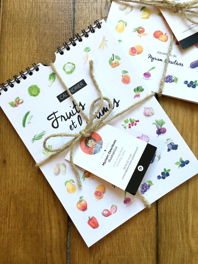 May include: A white calendar with a spiral binding featuring watercolor illustrations of fruits and vegetables. The calendar is titled "Fruits et L&eacute;gumes" and is tied with twine. A business card for Myriam Descl&egrave;ves, Illustratrice, is attached to the twine.