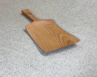 May include: A light brown wooden scoop with a flat, wide blade and a short, sturdy handle. The natural wood grain is visible across the surface of the scoop, which features a smooth, curved design that tapers from the handle to the wide edge.