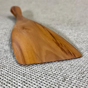 Könnte beinhalten: Ein Küchenspatel aus Holz mit glatter, hellbrauner Oberfläche und sichtbarer natürlicher Holzmaserung. Das Utensil verfügt über eine breite, flache Klinge, die in einen abgerundeten Griff übergeht und die organische Textur des Holzes zeigt.