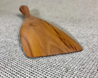 May include: A wooden kitchen spatula with a smooth, light brown finish and visible natural wood grain patterns. The utensil features a wide, flat blade that tapers into a rounded handle, showcasing the organic texture and warm color of the wood.