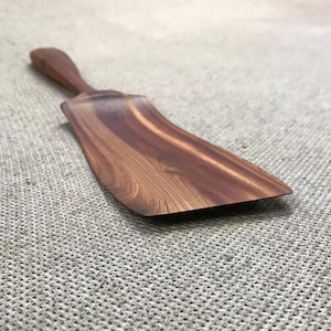 Könnte beinhalten: Ein Küchenspatel aus dunkelbraunem Holz mit sichtbarer natürlicher Maserung. Das Utensil verfügt über einen langen, schlanken Griff, der in eine breite, flache und leicht gebogene Klinge übergeht, die zum Anheben und Wenden von Speisen dient.