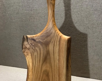 May include: A handcrafted wooden cutting board with a natural edge and a handle featuring a hole for hanging. The board is made of light brown wood with visible grain patterns. The board is approximately 12 inches long.