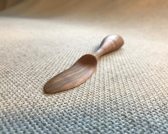 May include: A wooden butter knife with a curved handle and a pointed tip. The knife is made of dark wood with a natural grain pattern.