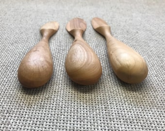 May include: Three wooden spoons with a teardrop-shaped bowl and a rounded handle. The spoons are made of light-colored wood and have a smooth, polished finish. They are arranged on a textured, neutral-colored surface.