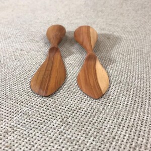 Peut inclure: Deux petits épandeurs en bois avec une finition lisse et polie et des motifs de grain naturel visibles. Chaque ustensile présente une lame large et arrondie et un manche effilé, mettant en valeur des tons de bois brun clair et foncé.