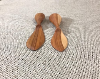 May include: Two small wooden spreaders with a smooth, polished finish and visible natural grain patterns. Each utensil features a wide, rounded blade and a slender, tapered handle, showcasing light and dark brown wood tones in a symmetrical design.