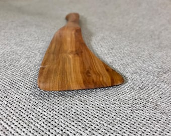 May include: A wooden spatula with a smooth, polished finish and visible natural grain patterns. The utensil features a wide, flat blade that tapers toward a narrow handle, showcasing the warm brown tones and organic texture of the wood grain.