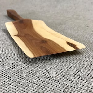 May include: A wooden serving spatula featuring a flat, rectangular blade with a natural live edge and a tapered handle. The wood displays a distinct contrast between dark brown heartwood in the center and lighter cream-colored sapwood on the sides.