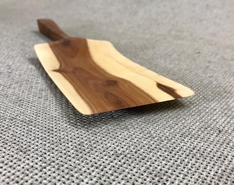 May include: A wooden serving spatula featuring a flat, rectangular blade with a natural live edge and a tapered handle. The wood displays a distinct contrast between dark brown heartwood in the center and lighter cream-colored sapwood on the sides.