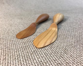 May include: Two wooden butter spreaders with curved blades and rounded handles. One is a light brown color, and the other is a darker brown. The spreaders are made of wood and are designed for spreading butter or other soft foods.