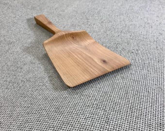 May include: A light brown wooden scoop with a smooth, polished surface and a visible natural wood grain pattern. The scoop features a wide, flat blade that tapers into a sturdy, integrated handle, showcasing the organic texture of the wood.