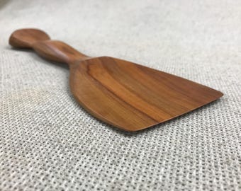 May include: A medium-brown wooden spatula with a smooth, polished finish and a distinct grain pattern. The utensil features a wide, flat, rectangular blade that tapers into a contoured, rounded handle, showcasing a natural wood texture throughout.