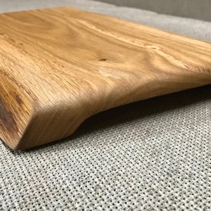 Può includere: Un tagliere rettangolare in legno con finitura a venature naturali. La tavola ha un manico curvo su un lato e un'area scura e strutturata sul bordo, che aggiunge un tocco rustico. La tavola è realizzata in legno marrone chiaro.