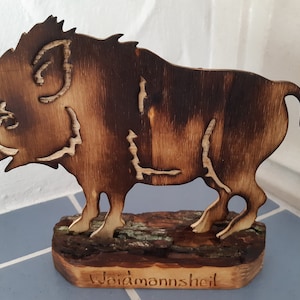 Könnte beinhalten: Eine Holzschnitzerei eines Wildschweins mit dunkelbrauner Oberfläche. Das Wildschwein steht auf einem Holzsockel mit dem eingravierten Text "Waidmannsheil".