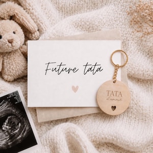 Peut inclure: Image aux tons neutres avec un lapin en peluche, une carte avec "Future tata" et un cœur, un porte-clés "TATA d'amour", et une échographie. L'ensemble est disposé sur une couverture texturée, évoquant une annonce de grossesse.