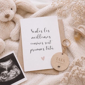 May include: A baby announcement card with the text "Seules les meilleures cousines sont promues tata" and a small pink heart. A wooden keychain with the text "TATA d'amour" hangs next to the card. A baby ultrasound image and a pacifier are also visible.